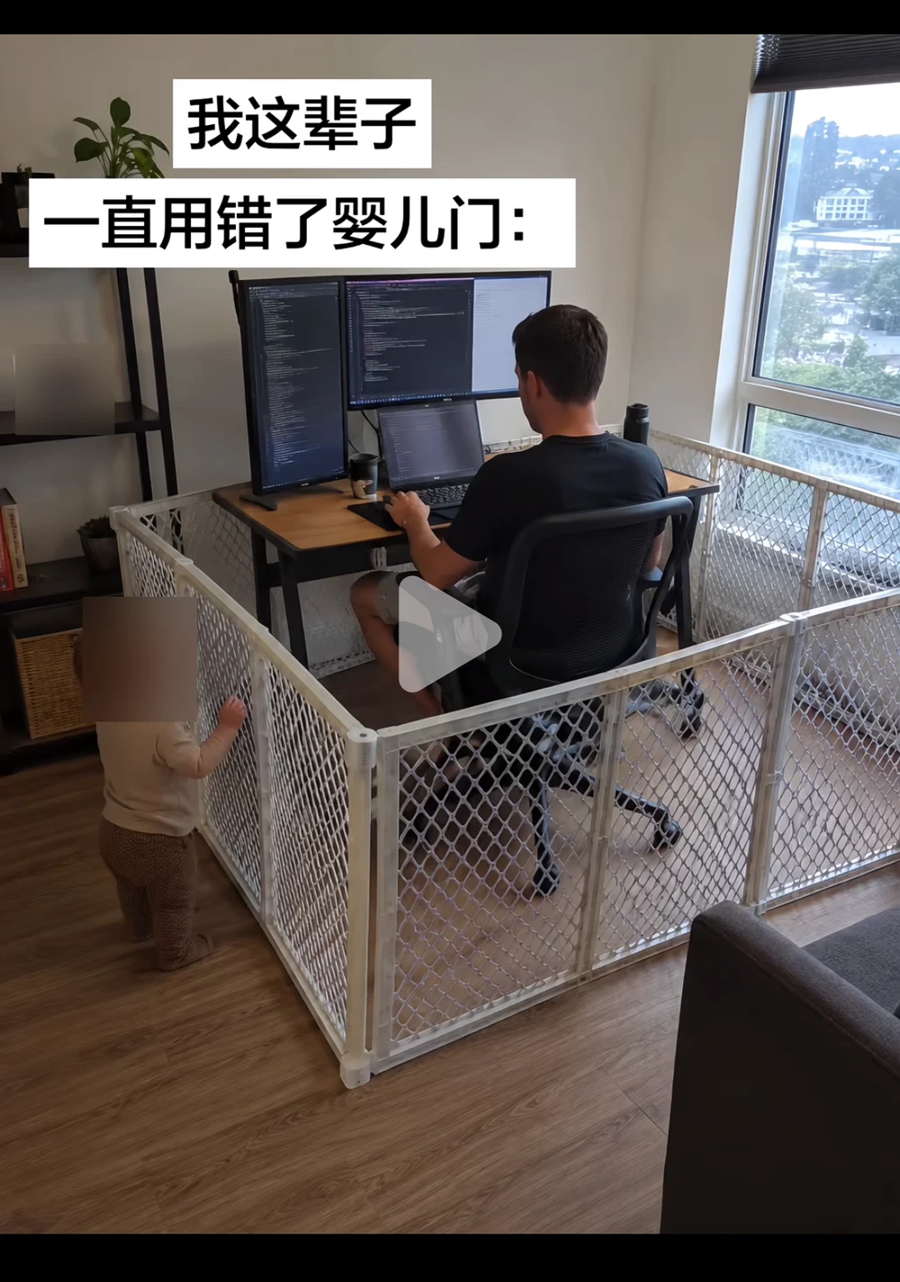 A father works at a three-monitor desk inside a white baby playpen while his toddler stands outside it, hands on the mesh