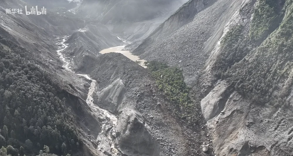 Aerial view of a Himalayan valley filled with grey debris after the mudslide, a muddy barrier lake pooled at its center, mist over the ridgelines